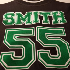 Może przedstawiać: Czarna i zielona kurtka bejsbolowa z nazwiskiem "SMITH" i numerem "55" na plecach.