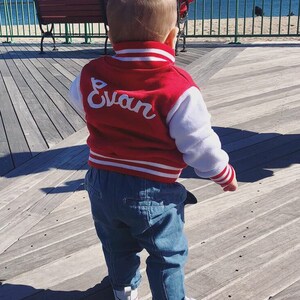 Może przedstawiać: Małe dziecko w czerwono-białej kurtce bejsbolowej z wyhaftowanym z tyłu imieniem "Evan". Kurtka ma białe rękawy i czerwone paski na mankietach i w pasie. Dziecko nosi niebieskie dżinsy i białe buty sportowe.