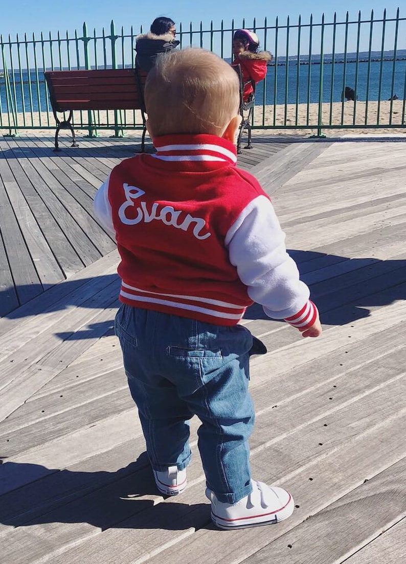 Może przedstawiać: Małe dziecko w czerwono-białej kurtce bejsbolowej z wyhaftowanym z tyłu imieniem "Evan". Kurtka ma białe rękawy i czerwone paski na mankietach i w pasie. Dziecko nosi niebieskie dżinsy i białe buty sportowe.