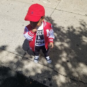 May include: A young child wearing a red baseball cap, a red and white varsity jacket with the letter "N" on the front, a white t-shirt with the text "Missed a Don't Karma" printed on it, and black trousers. The child is standing on a pavement.