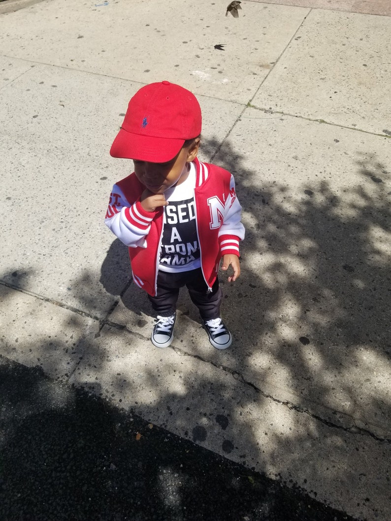 May include: A young child wearing a red baseball cap, a red and white varsity jacket with the letter "N" on the front, a white t-shirt with the text "Missed a Don't Karma" printed on it, and black trousers. The child is standing on a pavement.