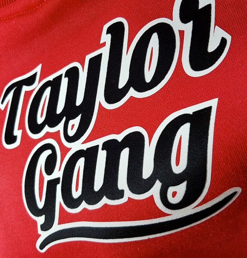 Pode incluir: Camisola vermelha com texto preto que l&ecirc; "Taylor Gang" em um tipo de letra estilizado.