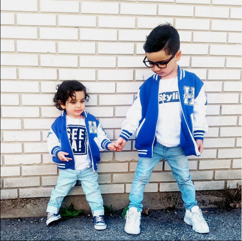 Może przedstawiać: Dw&oacute;ch młodych chłopc&oacute;w w niebieskich i białych kurtkach bejsbolowych. Chłopiec po lewej stronie ma na sobie białą koszulkę z nadrukiem "See Ya!" Chłopiec po prawej stronie ma na sobie białą koszulkę z nadrukiem "Styli".