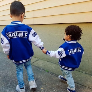 Może przedstawiać: Dw&oacute;ch młodych chłopc&oacute;w w niebieskich i białych kurtkach bejsbolowych z napisem "Ezelddine" na plecach. Chłopcy trzymają się za ręce i idą chodnikiem.