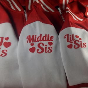 May include: Red and white letterman jackets with the phrases "Big Sis", "Middle Sis", and "Lil Sis" printed on the sleeves in red. The jackets have white sleeves and red bodies with white stripes on the collar and cuffs.