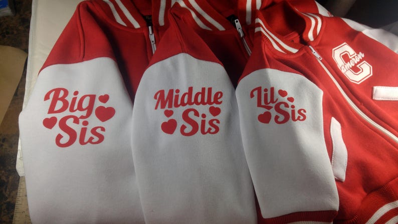 May include: Red and white letterman jackets with the phrases "Big Sis", "Middle Sis", and "Lil Sis" printed on the sleeves in red. The jackets have white sleeves and red bodies with white stripes on the collar and cuffs.