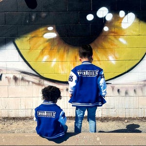 Może przedstawiać: Dw&oacute;jka dzieci w niebieskich i białych kurtkach bejsbolowych z napisem "Exelddine" na plecach stoi przed ceglaną ścianą z namalowanym na niej dużym okiem.