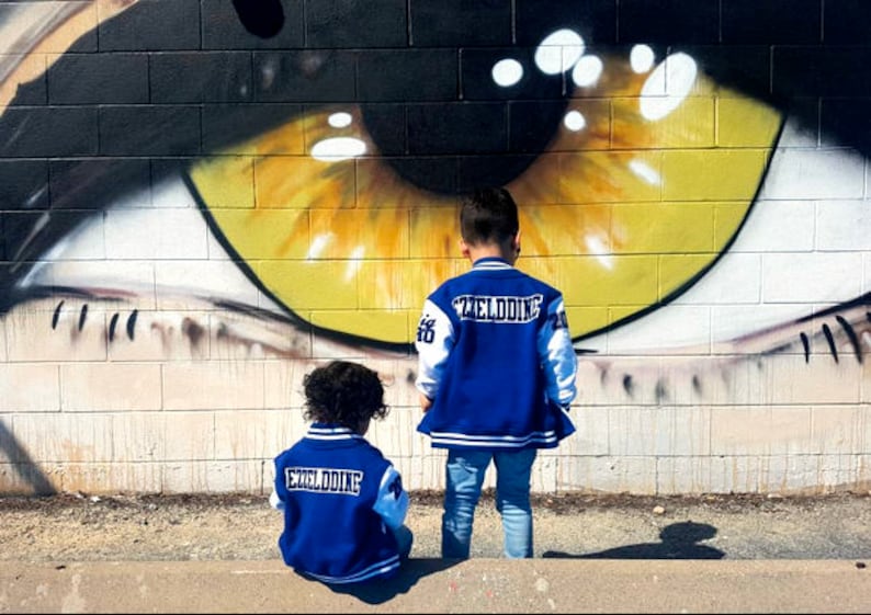 Może przedstawiać: Dw&oacute;jka dzieci w niebieskich i białych kurtkach bejsbolowych z napisem "Exelddine" na plecach stoi przed ceglaną ścianą z namalowanym na niej dużym okiem.