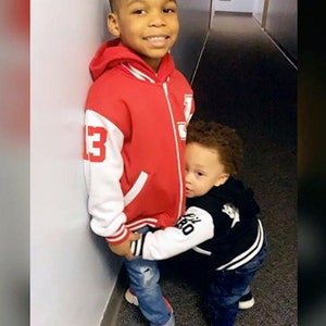 May include: Two young boys, one wearing a red and white baseball jacket and the other wearing a black and white baseball jacket, are standing in a hallway. The boy in the red jacket is smiling and the boy in the black jacket is looking at the camera.
