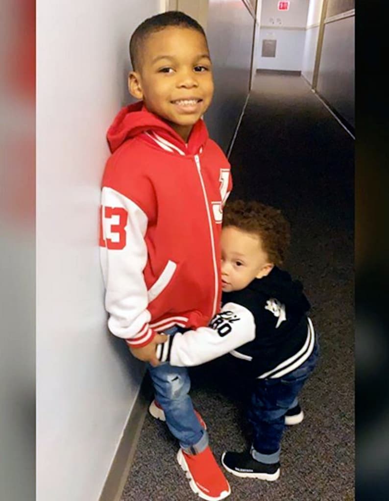May include: Two young boys, one wearing a red and white baseball jacket and the other wearing a black and white baseball jacket, are standing in a hallway. The boy in the red jacket is smiling and the boy in the black jacket is looking at the camera.