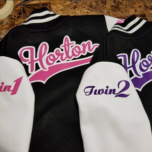 Pu&ograve; includere: Due giacche da baseball nere con maniche bianche. Le giacche hanno il nome "Horton" in corsivo rosa e viola sul retro. Le maniche hanno le parole "Twin 1" e "Twin 2" in corsivo rosa e viola.