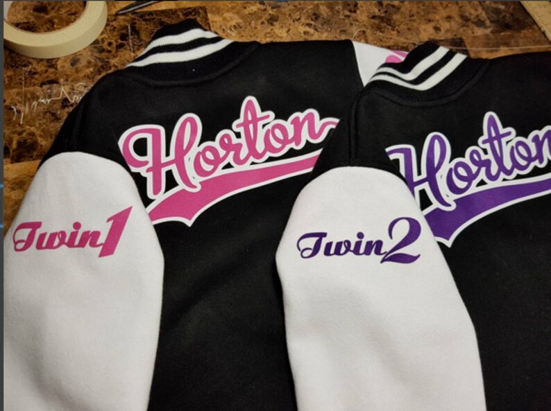 Pu&ograve; includere: Due giacche da baseball nere con maniche bianche. Le giacche hanno il nome "Horton" in corsivo rosa e viola sul retro. Le maniche hanno le parole "Twin 1" e "Twin 2" in corsivo rosa e viola.