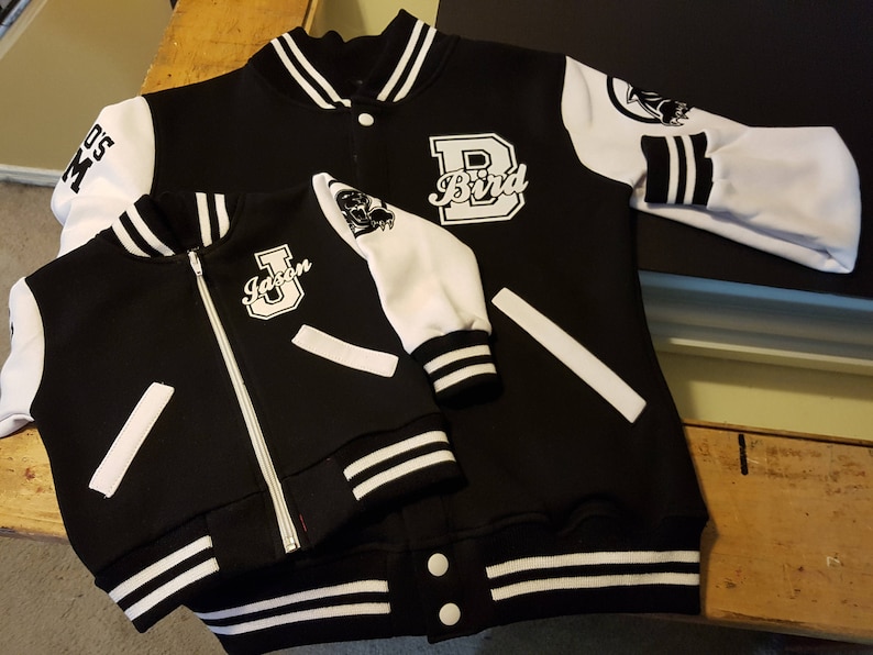 May include: Two black and white letterman jackets, one adult and one child size, are displayed. The adult jacket has "Bird" and a "B" on the front. The child's jacket has "Jason" on the front. Both have white sleeves.