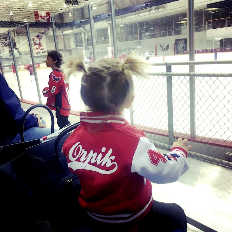 Pu&ograve; includere: Un bambino piccolo indossa una giacca da baseball rossa e bianca con il nome "Orpik" sul retro. Il bambino sta guardando una partita di hockey su una pista di ghiaccio.