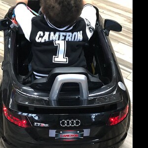 Pu&ograve; includere: Una giacca da baseball nera e bianca con il nome "CAMERON" e il numero "1" sul retro. La giacca &egrave; indossata da un bambino seduto in un'auto giocattolo nera che assomiglia a un'Audi.