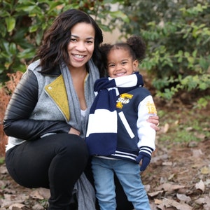 Może przedstawiać: Kobieta i młoda dziewczynka uśmiechają się do kamery. Kobieta ma na sobie szarą kurtkę ze sk&oacute;rzanymi rękawami i ż&oacute;łtym szalikiem. Dziewczynka ma na sobie niebiesko-białą kurtkę baseballową z niebiesko-białym szalikiem. Stoją przed zielonym krzewem.