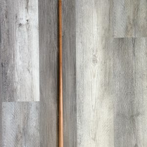 May include: A long, straight wooden pool cue with a smooth, light brown finish. The cue features a white ferrule at the tip, topped with a small, dark leather tip. The shaft tapers gradually from the thicker butt end toward the narrow tip end.