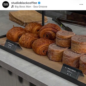May include: A display case containing golden-brown, flaky pain au chocolat pastries and round, cinnamon-dusted cinnamon rolls. Small black signs with white text label the items as Pain au Choc and Cinnamon Roll, with prices 3.8 and 4.3 respectively.