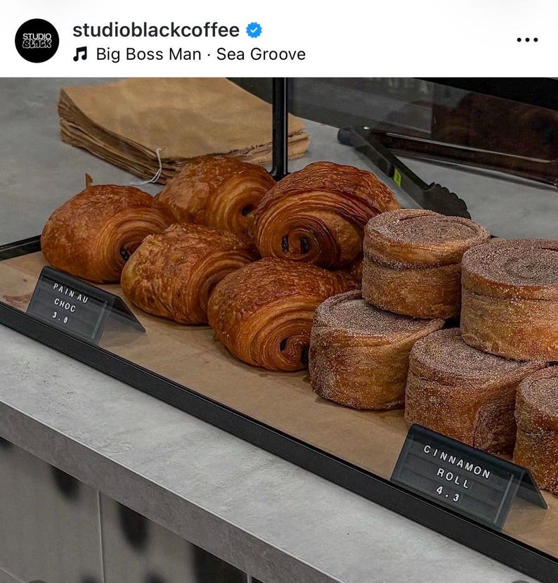 May include: A display case containing golden-brown, flaky pain au chocolat pastries and round, cinnamon-dusted cinnamon rolls. Small black signs with white text label the items as Pain au Choc and Cinnamon Roll, with prices 3.8 and 4.3 respectively.