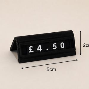 May include: A black triangular price tag display stand featuring white characters that read &pound; 4.50. The stand measures 5 cm in length and 2 cm in height as indicated by the black arrows and text labels printed on the image for reference.