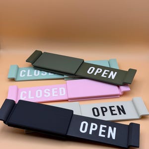 May include: A collection of rectangular sliding signs featuring the words OPEN or CLOSED in white or black lettering. The signs are available in various colors including olive green, light blue, pink, light gray, and black, with a sliding mechanism.