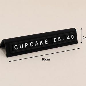May include: A black L-shaped price tag stand featuring white letter tiles that spell out CUPCAKE &pound; 5.40. The stand measures 10 cm in length and 2 cm in height as indicated by the black dimension arrows printed on the image.