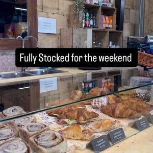 May include: A glass display case filled with various baked goods including cinnamon buns, bacon turnovers, and croissants. A black sign with white text reads "Fully Stocked for the weekend" above the pastries, with smaller labels identifying items.