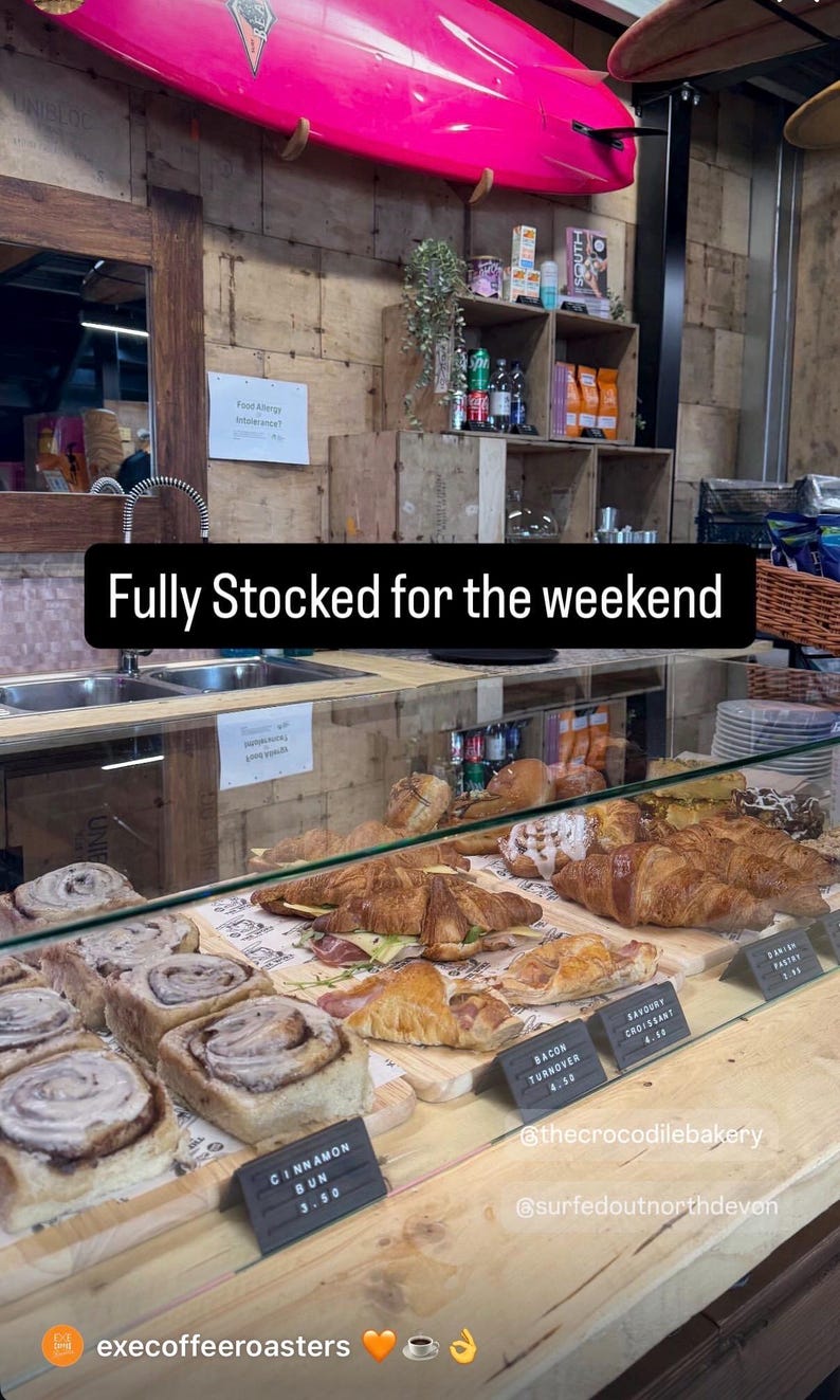May include: A glass display case filled with various baked goods including cinnamon buns, bacon turnovers, and croissants. A black sign with white text reads "Fully Stocked for the weekend" above the pastries, with smaller labels identifying items.
