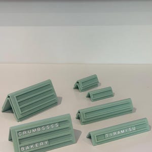 May include: A collection of sage green, V-shaped display stands with horizontal grooves for inserting small letter tiles. Two stands feature text: one reads CRUMBSSSS and BAKERY, while another reads TIRAMISU. The stands vary in length and height.