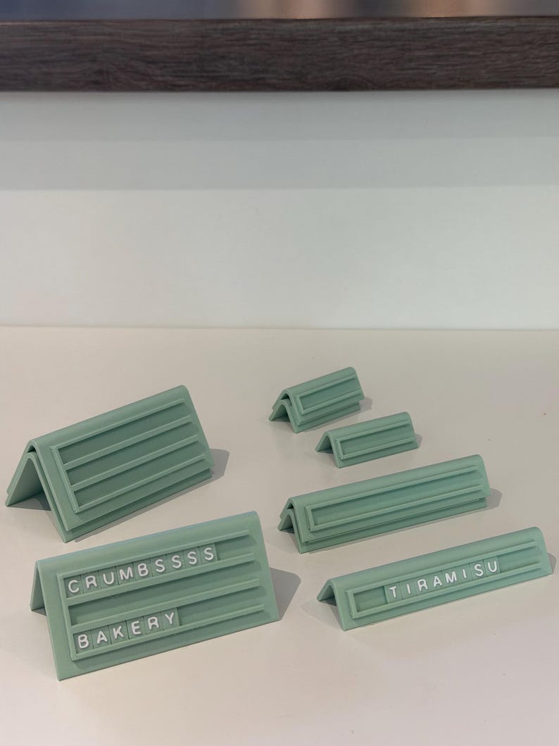 May include: A collection of sage green, V-shaped display stands with horizontal grooves for inserting small letter tiles. Two stands feature text: one reads CRUMBSSSS and BAKERY, while another reads TIRAMISU. The stands vary in length and height.