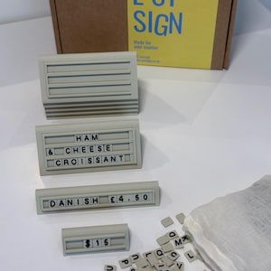 May include: A set of four light gray plastic sign holders with horizontal grooves for inserting black letter tiles. One holder displays HAM CHEESE CROISSANT, one shows DANISH &pound;4.50, and one shows $15. A white fabric bag contains loose letter tiles.