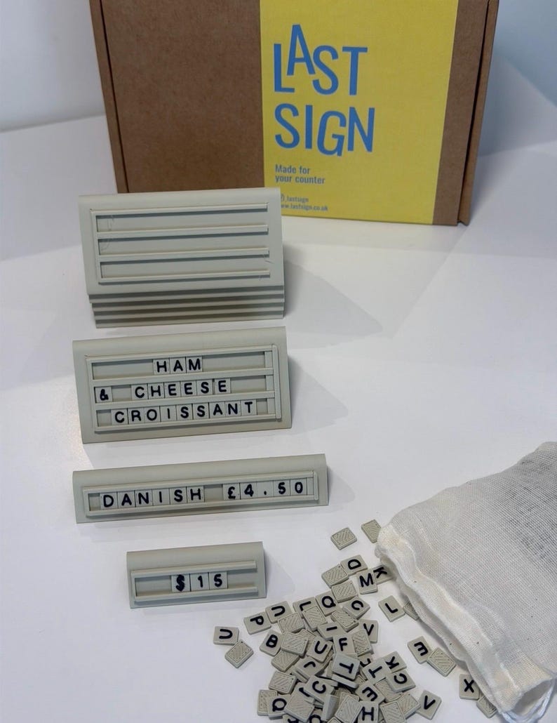 May include: A set of four light gray plastic sign holders with horizontal grooves for inserting black letter tiles. One holder displays HAM CHEESE CROISSANT, one shows DANISH &pound;4.50, and one shows $15. A white fabric bag contains loose letter tiles.