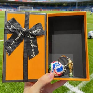 Puede incluir: Caja de regalo naranja abierta con una cinta negra que dice "love you forever". En el interior, un llavero de balón de fútbol y un llavero de trofeo dorado. La caja está sobre un campo verde.