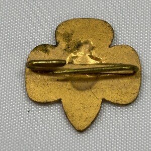 May include: A vintage gold-colored pin with a clover-like shape. The pin has a horizontal bar and a clasp on the back. The pin is likely a collectible item or a piece of jewelry.