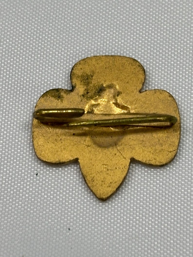 May include: A vintage gold-colored pin with a clover-like shape. The pin has a horizontal bar and a clasp on the back. The pin is likely a collectible item or a piece of jewelry.