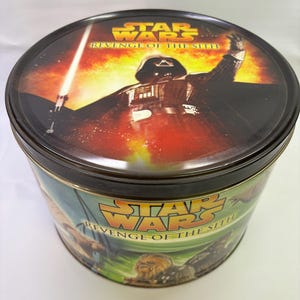 May include: A round, black tin featuring Star Wars: Revenge of the Sith imagery. The lid displays Darth Vader with a lightsaber against a fiery background. The sides show characters like Chewbacca.