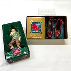 May include: A Wizard of Oz scarecrow ornament in a green box and a pair of red glittery slippers in a gold box with the text There's no place like Home for Christmas. The scarecrow wears a green shirt and brown pants with a gold hanging string.