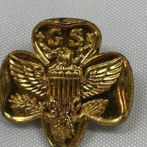 May include: A gold-colored Girl Scout pin in the shape of a trefoil, featuring an eagle with outstretched wings and the letters "G S". The pin has a detailed design with a shiny finish, and is likely made of metal.