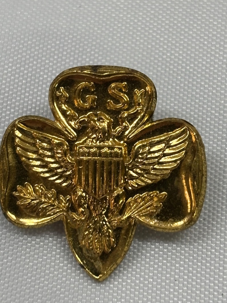May include: A gold-colored Girl Scout pin in the shape of a trefoil, featuring an eagle with outstretched wings and the letters "G S". The pin has a detailed design with a shiny finish, and is likely made of metal.