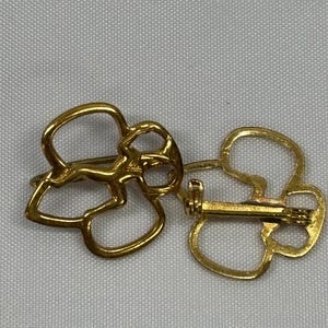 May include: Two gold-colored metal brooches, each shaped like a stylized four-leaf clover. The brooches are shown side-by-side, with one displaying the pin and clasp mechanism on the back. The brooches are on a light gray background.