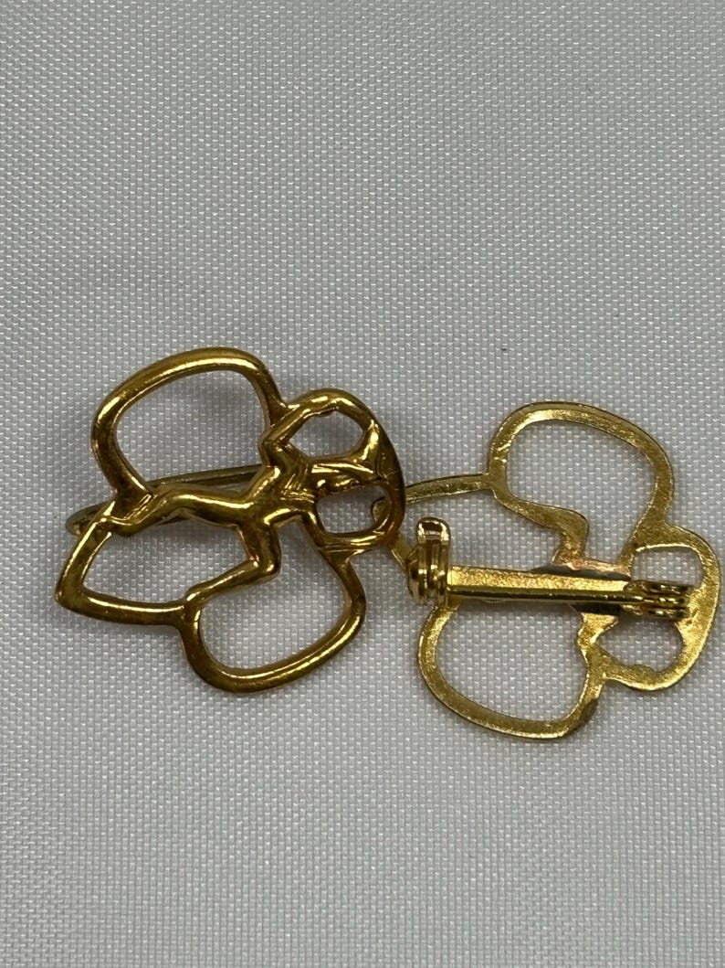 May include: Two gold-colored metal brooches, each shaped like a stylized four-leaf clover. The brooches are shown side-by-side, with one displaying the pin and clasp mechanism on the back. The brooches are on a light gray background.