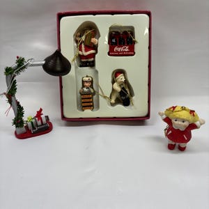 May include: A collection of holiday ornaments including a boxed set of four Coca-Cola themed figures, a Hershey's Kisses lamp ornament with small gift boxes, and a blonde doll ornament wearing a red dress with white trim and red bows.