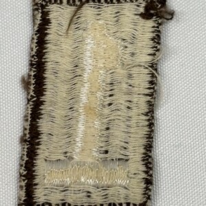 May include: A small, rectangular patch with a woven design. The patch has a cream-colored center with a vertical pattern and a dark brown border. Loose threads are visible at the top and edges, suggesting a handmade or vintage item.