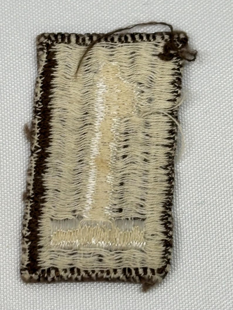 May include: A small, rectangular patch with a woven design. The patch has a cream-colored center with a vertical pattern and a dark brown border. Loose threads are visible at the top and edges, suggesting a handmade or vintage item.