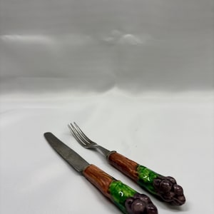 May include: A knife and fork set with handles designed to resemble asparagus. The handles are brown, green, and purple. The stainless steel knife and fork are placed on a white surface.