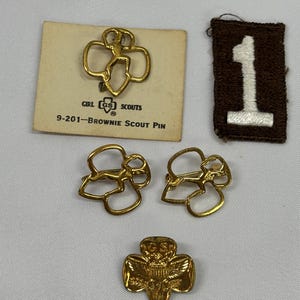 May include: A collection of Girl Scout memorabilia, including gold-colored pins and a brown patch with a white number one. One pin is on a card that reads "Girl Scouts" and "Brownie Scout Pin".