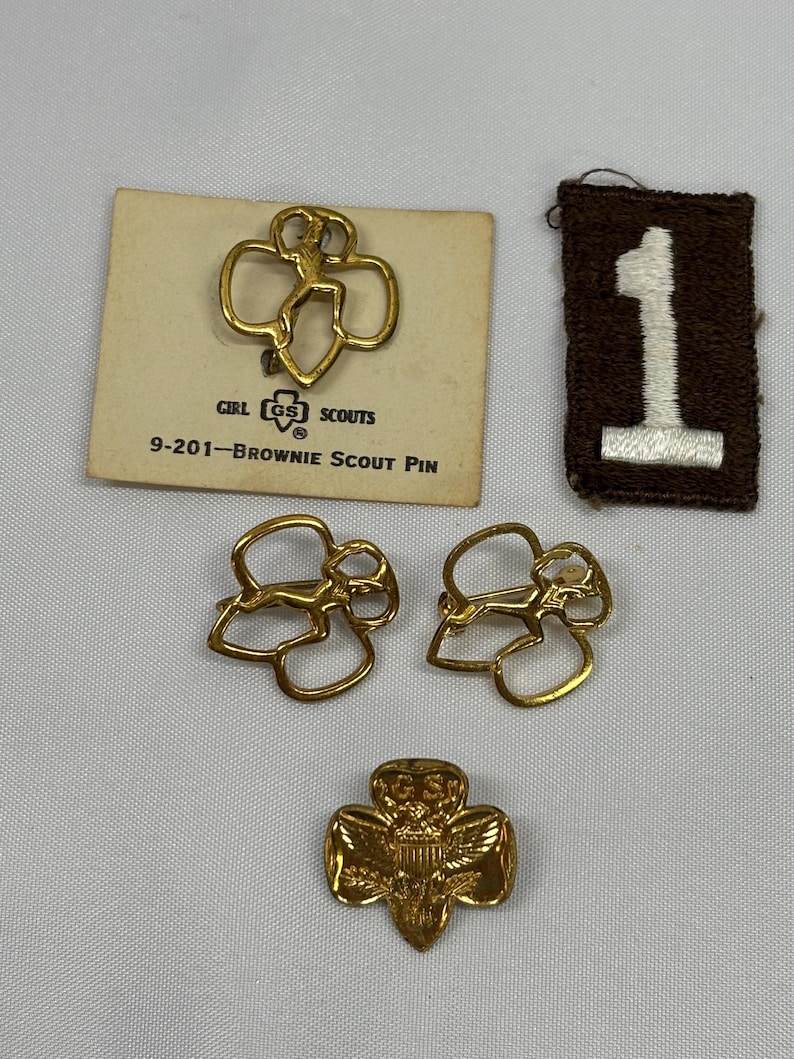 May include: A collection of Girl Scout memorabilia, including gold-colored pins and a brown patch with a white number one. One pin is on a card that reads "Girl Scouts" and "Brownie Scout Pin".