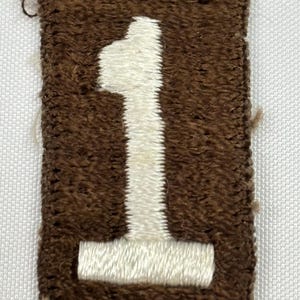 May include: A rectangular brown patch with a white embroidered number 1. The patch has a textured appearance, with the number prominently displayed in the center. The edges of the patch are also brown.