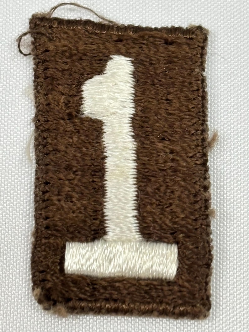 May include: A rectangular brown patch with a white embroidered number 1. The patch has a textured appearance, with the number prominently displayed in the center. The edges of the patch are also brown.