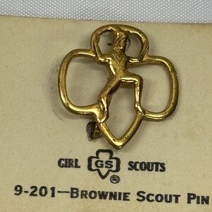 May include: A gold-colored Brownie Scout pin featuring a stylized figure within a trefoil shape. The pin is attached to a tan card with the Girl Scouts logo and the text "9-201-Brownie Scout Pin".